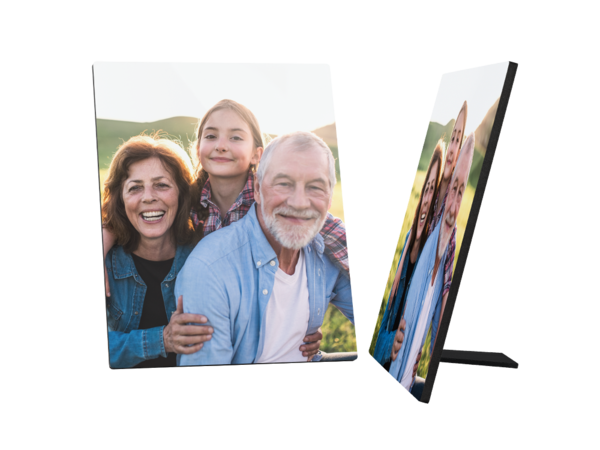 Ein Fotoaufsteller zeigt ein fröhliches Familienbild. Eine lächelnde ältere Frau mit braunen Haaren steht links. Hinter ihr steht ein junges Mädchen mit blonden Haaren in einem karierten Hemd und lächelt ebenfalls. Rechts von ihnen steht ein älterer Mann mit weißen Haaren und Bart, der auch fröhlich in die Kamera schaut und seine Arme um die beiden anderen gelegt hat.